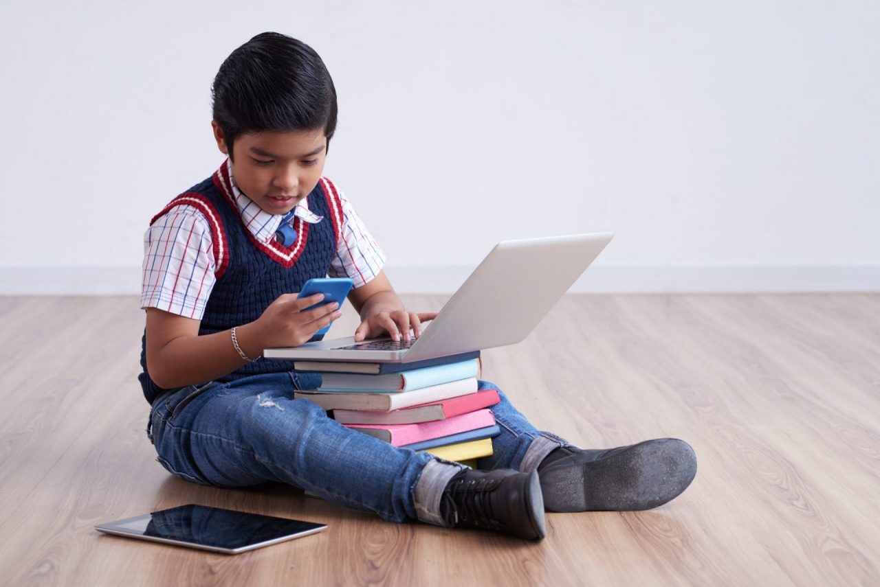 menino sentado no chão apoiando o computador em cima de vários livros e olhando o celular