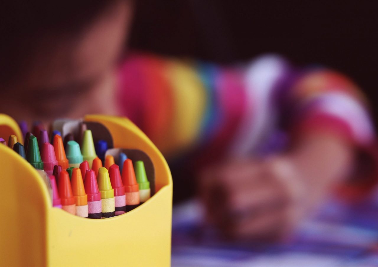 criança ao fundo pintando e a frente dela um porta lápis com muitos gizes de cera coloridos